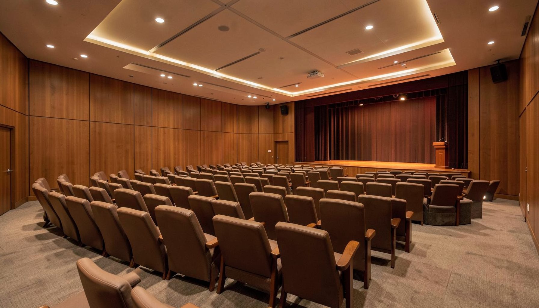 A wide conference room with rows of seating and a stage, warm wood tones and soft overhead lighting, professional atmosphere