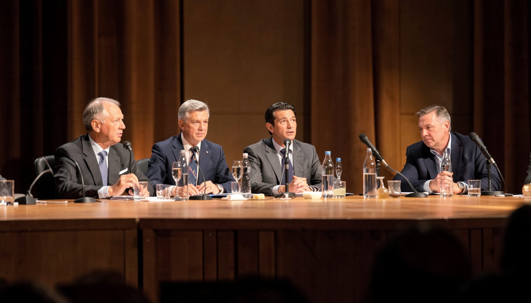 Panel discussion on stage with speakers at a long table, warm indoor lighting, professional setting